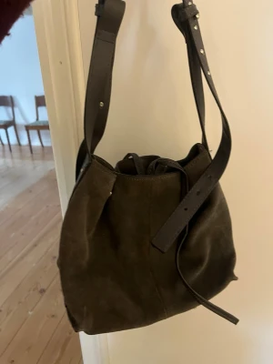 Grå väska - Snygg axelväska i mörkbrun mocka med justerbar axelrem. Väskan har en bucket-form och stängs med dragsko. Perfekt för dig som gillar en stilren och trendig look.