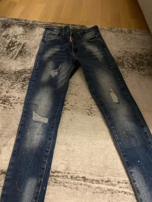 Mörkblå slitna skinny jeans Dsquared  - Säljer ett par mörkblå skinny jeans med slitningar och distressed detaljer på benen. Jeansen har en tvättad look och små färgstänk för en trendig, avslappnad stil. Perfekta för dig som gillar streetwear och vill ha en cool vibe.