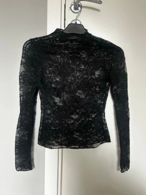 Svart spetsblus med lång ärm - Säljer en svart, transparent spetsblus med lång ärm och hög hals. Blusen har ett blommigt spetsmönster som ger en elegant och edgy vibe. Perfekt att styla lager på lager eller till fest💕