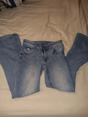 Ljusblå bootcut jeans med strass - Säljer ett par ljusblå bootcut jeans med strassdetaljer på framfickorna och en snygg dekoration i strass på ena bakfickan. Jeansen har klassisk femficksdesign och är tillverkade i denim med lätt tvättad look. Perfekta för dig som gillar lite extra bling i vardagen.