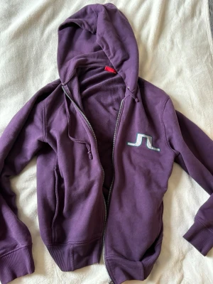Lila vintage hoodie från J.Lindeberg - Säljer en lila vintage hoodie från J.Lindeberg med dragkedja och huva. Tröjan har ett broderat vitt och grönt J.Lindeberg-logo på bröstet, ribbade muddar och fickor framtill i storlek S