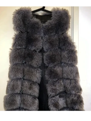 Grå fluffig pälsväst - Snygg och trendig grå pälsväst med stora, mjuka pälspartier i blockmönster. Västen är ärmlös och har en rak passform som ger en cool och lyxig känsla. Perfekt att slänga över en hoodie eller tröja för extra stilpoäng.