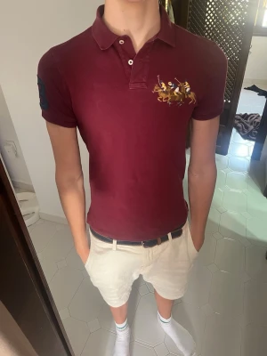 Vinröd vintage piké från Ralph Lauren - Snygg vinröd pikétröja från Ralph Lauren med klassisk krage, knappar och broderat polospel-motiv på bröstet. Tröjan har korta ärmar och är tillverkad i mjuk bomull som känns skön mot huden. Perfekt för en stilren och avslappnad look.