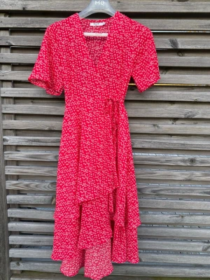 Jättefin omlottklänning från NAKD (34)  - Säljer nu denna fantastiska röda sommarklänning med ett sött vitt blommönster. Klänningen har en smickrande omlottmodell med knytning i midjan och fina volangdetaljer i nederkanten som ger ett härligt fall. • Märke: NA-KD • Storlek: 34 • Skick: Som ny! Endast använd vid ett enstaka tillfälle (studentfirande) • Passform: Klassisk omlott. 