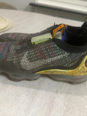 Nike Air Vapormax Flyknit multicolor - Säljer ett par Nike Air Vapormax Flyknit sneakers med svart bas och färgglada detaljer i stickat material. Skorna har en genomskinlig sula, elastisk ovandel och smarta detaljer som pull tabs och en grön spänne på ovansidan. Perfekta för dig som gillar unika och bekväma sneakers.