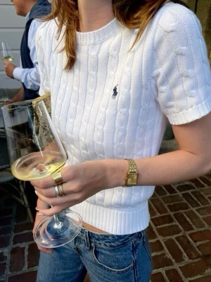 Vit kabelstickad topp från Polo Ralph Lauren - Kabelstickad vit topp från Polo Ralph Lauren med korta ärmar och rund hals. Klassisk broderad logga på bröstet. Tillverkad i mjuk bomull som ger en skön och stilren look. Perfekt till jeans eller kjol för en clean outfit.