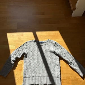 Grå kabelstickad tröja Polo Ralph Lauren - Kabelstickad grå tröja från Polo Ralph Lauren med rund halsringning och diskret broderad logga på bröstet. Tröjan har långa ärmar och breda ribbade muddar vid ärmslut och nederkant. Perfekt för lager-på-lager under höst och vinter.