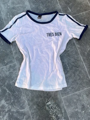 Vit t-shirt Très Bien Gina Tricot - Vit t-shirt från Gina Tricot med mörkblå detaljer vid hals och ärmslut med trycket 'TRÈS BIEN' på bröstet . Klassisk passform och korta ärmar, perfekt till jeans eller kjol.