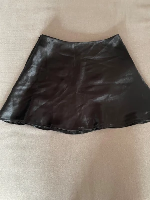 Svart glansig satinkjol - Snygg svart kjol i glansigt satinmaterial med lätt utställd A-linjeform. Kjolen har en diskret dragkedja i sidan för enkel på- och avtagning. Perfekt för dig som gillar en stilren och trendig look.