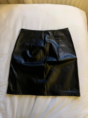 Svart skinnkjol i kort modell - Snygg svart kort kjol i skinnimitation med diskreta sömdetaljer. Kjolen har två dekorativa fickor bak och en stilren look som passar till många olika outfits. Köptes för år sedan men har bara blivit liggandes, från Gina tricot.