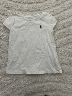 Vit t-shirt från Polo Ralph Lauren - Klassisk vit t-shirt från Polo Ralph Lauren med den ikoniska mörkblå loggan broderad på bröstet. T-shirten har rund halsringning och korta ärmar. Tillverkad i mjuk bomull som känns skön mot huden. Perfekt basplagg till garderoben.