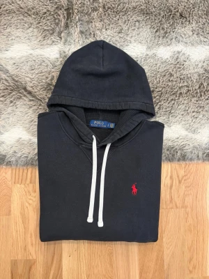 Svart Hoodie Från Polo Ralph Lauren - Hej! 👋 Säljer en stilren svart Polo Ralph Lauren hoodie i storlek S (Herr). Den har den klassiska röda pony-loggan broderad på bröstet 🐎 Tröjan är i bra skick och har den sköna mjuka fleece-insidan kvar ✨ Det kan förekomma en naturlig, lätt solblekning på vissa ställen, vilket ger den en snygg vintage-känsla 👕 Sitter perfekt (Modell: 177cm). Modellen saknar QR-kod men alla tvättlappar sitter kvar ✅ Ett oumbärligt basplagg som passar till allt! Fraktar snabbt 🚛📦💬💯 