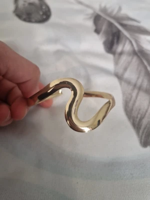 Guldigt vågformat armband - Snyggt armband i guldfärgad metall med unik vågformad design. Öppet avslut som gör det enkelt att ta på och av. Perfekt för dig som gillar trendiga och stilrena accessoarer med en twist.