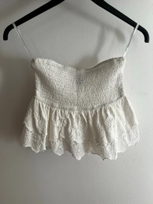 Vit broderad tubtop med volang - Söt vit tubtop med smockad överdel. Toppen har broderade blommor och dubbla volanger nedtill för en romantisk vibe. Perfekt till sommaren och lätt att matcha med jeans eller kjol.