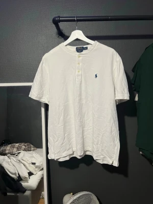 Vit t-shirt från Polo Ralph Lauren - Vit t-shirt från Polo Ralph Lauren med korta ärmar och knappdetaljer vid halsen. Klassisk blå logga broderad på bröstet. Tillverkad i mjuk bomull och har en avslappnad passform som passar perfekt till jeans eller shorts.