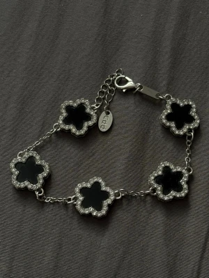 Silverarmband med svarta blommor - Snyggt armband i silverfärg med fem blommor i svart och gnistrande stenar runt varje blomma. Länken är tunn och har justerbar längd med karbinlås. Perfekt för dig som gillar stilrena och eleganta accessoarer med lite extra bling.