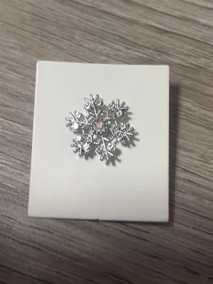 Snöflingeformad brosch i silverfärg - Säljer en snygg brosch i form av en snöflinga i silverfärg med små glittrande stenar och en skimrande sten i mitten. Perfekt att fästa på jackan eller väskan för en vintrig touch. Broschen har nål och säkerhetslås på baksidan.