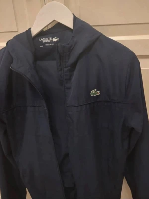 Mörkblå vindjacka från Lacoste - Snygg mörkblå vindjacka från Lacoste Sport med dragkedja och huva. Jackan har det klassiska gröna krokodilmärket på bröstet och är tillverkad i ett lätt material som passar perfekt för blåsiga dagar. Enkel och stilren design som funkar till det mesta. Tar även imot byten 