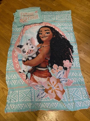 Moana påslakanset med Pua - Fint påslakanset med motiv från Disneyfilmen Moana. Setet har turkosa och rosa färger med mönster och texten 'Explore Oceania' på örngottet. Stort tryck av Moana som kramar grisen Pua, samt blommor och blad. Perfekt för dig som älskar Disney och vill ha ett färgglatt sovrum.