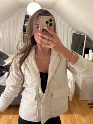 Vit quiltad jacka med fickor - Stilren vit quiltad jacka med stora fickor framtill och knappstängning. Jackan har lång ärm och en klassisk krage, perfekt för dig som gillar en clean och modern look. Passar till många olika outfits och är lätt att matcha.