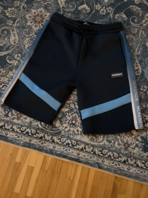 Svarta shorts från McKenzie med blå detaljer - Säljer ett par svarta McKenzie shorts med ljusblåa ränder och logga på benet. Shortsen har snörning i midjan och breda sidopaneler med McKenzie-tryck. Perfekta för sommaren och sportiga outfits.