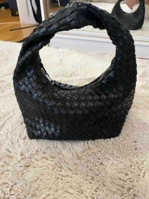 Svart flätad handväska i läderstil - Snygg svart handväska med flätad design och rundat handtag. Väskan har en unik form och är tillverkad i material som liknar läder. Perfekt för dig som vill ha en trendig accessoar med cool struktur och modern look.