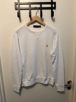 Vit sweatshirt från Polo Ralph Lauren - Klassisk vit sweatshirt från Polo Ralph Lauren med rund halsringning och den ikoniska broderade loggan i orange på bröstet. Tröjan har ribbade muddar vid ärmslut och nederkant, samt en relaxed passform som passar perfekt till jeans eller sweatpants.