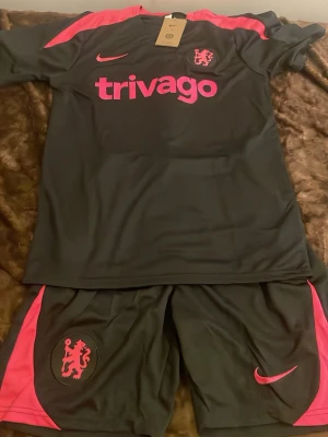 Chelsea träningsset svart/rosa Nike - Säljer ett Chelsea träningsset från Nike i svart med rosa detaljer. Setet består av en t-shirt och shorts med klubbmärke, Nike-logga och trivago-tryck. Materialet är lätt och andas, perfekt för träning eller match. Snygg kontrast mellan svart och rosa.