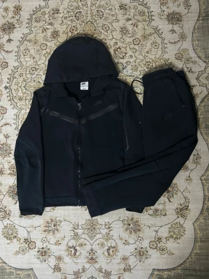 Svart Nike hoodie med dragkedja - Säljer en svart hoodie från Nike med dragkedja framtill och huva. Hoodien har diskreta svarta detaljer över bröstet och Nike-logga på vänster sida. Tillverkad i mjukt bomullsmaterial som känns skönt mot huden. Perfekt för en avslappnad och sportig stil.