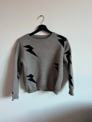 Grå stickad tröja med blixtmotiv - Säljer en grå/beige stickad tröja med svarta blixtar som detaljer. Tröjan har rund halsringning, långa ärmar och ribbade muddar. Perfekt för dig som gillar en cool och avslappnad stil. Materialet känns mjukt och bekvämt. Den har blixtar som mönster ⚡️⚡️
