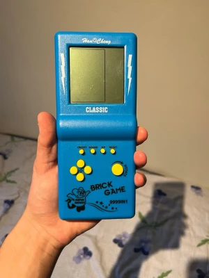 Blå Brick Game handhållen konsol - Retro handhållen spelkonsol i blå plast från Hua Cheng med svart text och gula knappar. Skärmen är monokrom och konsolen har klassisk Tetris-design med texten 'BRICK GAME 9999 IN 1' på framsidan. Perfekt för nostalgiska spelstunder.