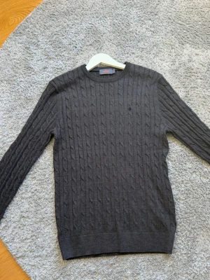 Mörkgrå kabelstickad tröja Morris - Snygg mörkgrå kabelstickad tröja från Morris med rund halsringning och diskret logga broderad på bröstet. Tröjan har långa ärmar och ribbade muddar vid ärmslut och nederkant. Använd 1 gång.