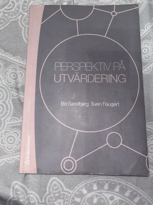 Perspektiv på utvärdering bok - Kurslitteratur med titeln 'Perspektiv på utvärdering' av Bo Sandberg och Sven Faugert. Boken har ett matt omslag i grå och rosa toner med grafiskt cirkelmönster. Perfekt för dig som pluggar utvärderingsmetodik eller samhällsvetenskap.