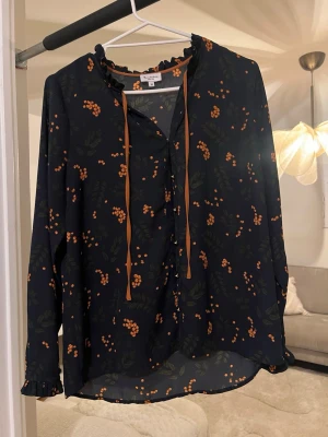 Svart blus med orange blommönster - Snygg mörkblå blus från Twist & Tango med orange blommönster och tunna knytband vid halsen. Blusen har långa ärmar med små volangdetaljer vid ärmslut och halsringning. Tillverkad i ett lätt och luftigt material som ger en skön känsla.