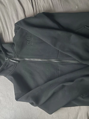 Svart hoodie från C.P. Company - Snygg svart hoodie från C.P. Company med dragkedja framtill och diskret logga broderad på bröstet. Klassisk passform med huva och mjukt material som känns skönt mot huden. Perfekt för en avslappnad streetwear-look.