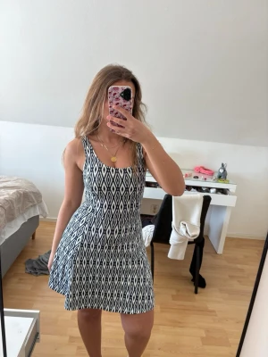 Kort ärmlös klänning med mönster - Säljer en kort, ärmlös klänning med  grafiskt mönster. Klänningen har en rundad urringning och sitter figurnära upptill med en lätt utställd kjol. Perfekt för varma dagar och enkel att styla med accessoarer. Aldrig använd!