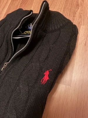 Svart kabelstickad tröja Polo Ralph Lauren - Svart kabelstickad tröja från Polo Ralph Lauren med dragkedja och hög krage. Tröjan har det klassiska röda Polo-logotypbroderiet på bröstet och ribbade muddar. Perfekt för dig som gillar stilrena och tidlösa plagg.