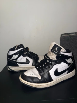 Air Jordan 1 Mid svart/vit - Säljer ett par Air Jordan 1 Mid sneakers i svart och vitt läder med klassisk Swoosh på sidan och Jordan Wings-logga vid ankeln. Skorna har svart snörning och vit mellansula. Perfekta för dig som gillar streetwear och ikonisk design.