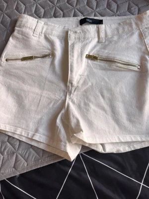 Vita shorts med dragkedjor från Ellos - Snygga vita shorts från Ellos med raka ben och två guldfärgade dragkedjor framtill. Klassisk femficksmodell med bälteshällor och bakfickor. Perfekta för sommardagar och lätta att matcha med olika toppar.