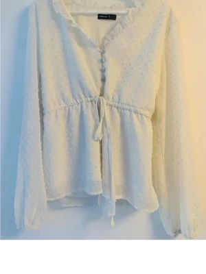 Vit blus med knappar från Shein - Söt vit blus från Shein med diskreta prickar i tyget, volangkant vid halsen och knappar framtill. Blusen har knytband i midjan och långa, luftiga ärmar. Perfekt till jeans eller kjol för en romantisk look.