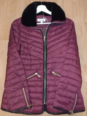 Vinröd pufferjacka från Viveca N - Stilren vinröd pufferjacka från Viveca N med svarta detaljer och mjuk krage i fuskpäls. Jackan har guldfärgade dragkedjor på fickor och ärmslut samt quiltad design. Perfekt för kyliga dagar och ger en snygg siluett.