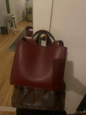 Vinröd handväska i skinnimitation - Säljer en stilren vinröd handväska i skinnimitation med både handtag och axelrem. Väskan har en fyrkantig form och minimalistisk design utan synliga loggor eller detaljer. Perfekt för dig som gillar enkel och modern stil.