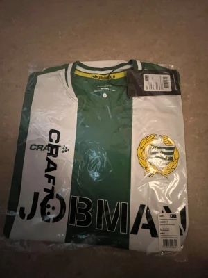 Hammarby matchtröja grön/vit Craft - Säljer en officiell äkta Hammarby matchtröja från Craft i grönt och vitt med klubbmärke på bröstet och JOBMAN Workwear som sponsortryck. Tröjan har klassiska breda ränder, rund hals och är tillverkad i ett lätt, funktionellt material perfekt för fotboll.