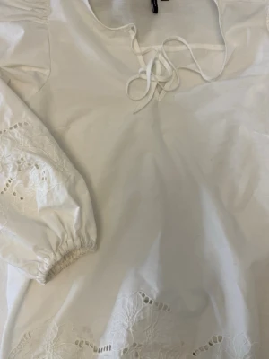 Vit blus med broderade detaljer - Söt vit blus med långa puffärmar och broderade blommor på ärmar och nederkant. Blusen har knytning vid halsen och är gjord i ett lätt bomullstyg. Perfekt för en romantisk och avslappnad stil. Aldrig använd. 