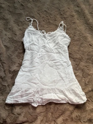 Vit linnetopp från Pull&Bear - Supersöt vit linnetopp från Pull&Bear med tunna axelband och volangkant nertill. Toppen är ärmlös och har en lätt och luftig känsla, perfekt för varma dagar. Tillverkad i bomull och linne för en fräsch sommarlook.