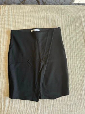 Svart omlottkjol från Gina Tricot - Säljer en svart kort omlottkjol från Gina Tricot. Kjolen har en enkel och stilren design med omlottdetalj framtill. 