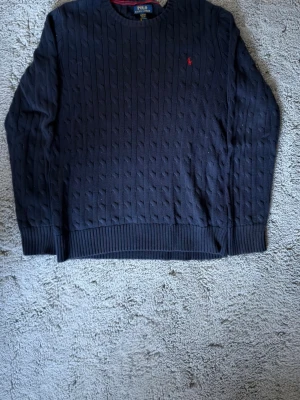 Polo Ralph lauren tröja - Kabelstickad mörkblå tröja från Polo Ralph Lauren med rund halsringning och diskret röd logga broderad på bröstet. Tröjan har ribbade muddar vid ärmslut och nederkant. Perfekt för dig som gillar klassisk och stilren design.