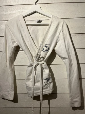 Vit omlott tröja från Better Bodies - Mysig vit omlott tröja från Better Bodies med broderad logga på bröstet och texten 'Wellness' på fickan. Modellen har långa ärmar, knytskärp i midjan och två fickor framtill. Perfekt för att chilla hemma eller till yoga klassen . Strl L.