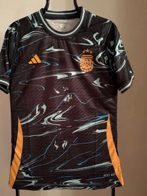 Argentina fotbollströja svart/blå - Snygg svart och blå Argentina fotbollströja från Adidas med orange detaljer och tryck. Framsidan har AFA-logga och Adidas-märke, baksidan har stort tryck med texten 'Campeones de América 2024' och motiv av en fotboll. Tillverkad i lätt och ventilerande material.