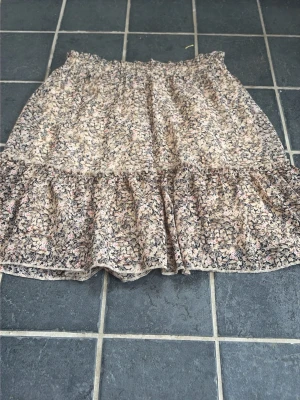 Blommig volangkjol i beige och rosa - Supersöt kortkjol med blommigt mönster i beige, rosa och grå toner. Kjolen har volang nedtill och resår i midjan för en bekväm passform. Perfekt för varma dagar och enkel att matcha med en enkel topp.